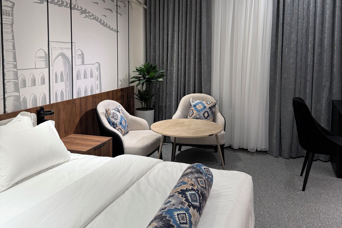 Habitación de hotel moderna con una cama grande, una pequeña mesa redonda, dos sillones, un escritorio y un amplio ventanal con cortinas.
