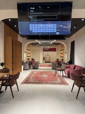 Un hall d’hôtel moderne doté d’un grand téléviseur à écran plat affichant une page de résultats de recherche Google, d’un espace salon avec des canapés et des fauteuils, d’un grand tapis à motifs et d’une étagère en bois.