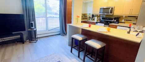 Una cocina moderna con una isla central, taburetes de bar, un televisor de pantalla plana y un ventanal corredero que da a un balcón.