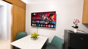Et moderne køkken med et fladskærms-tv, der viser en film, et spisebord med stole, et køleskab og en potteplante.