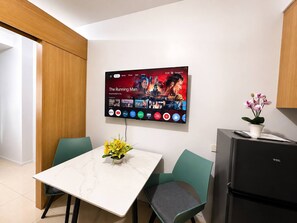 A modern kitchen with a flat-screen TV displaying a movie, a dining table with chairs, a refrigerator, and a potted plant. - Perky 1-bedroom Apartment in Vibrant Pasay City (Pasay)