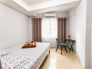 A compact hotel room with a bed, a small table with a vase of flowers, a chair, and an air conditioning unit. - Perky 1-bedroom Apartment in Vibrant Pasay City (Pasay)