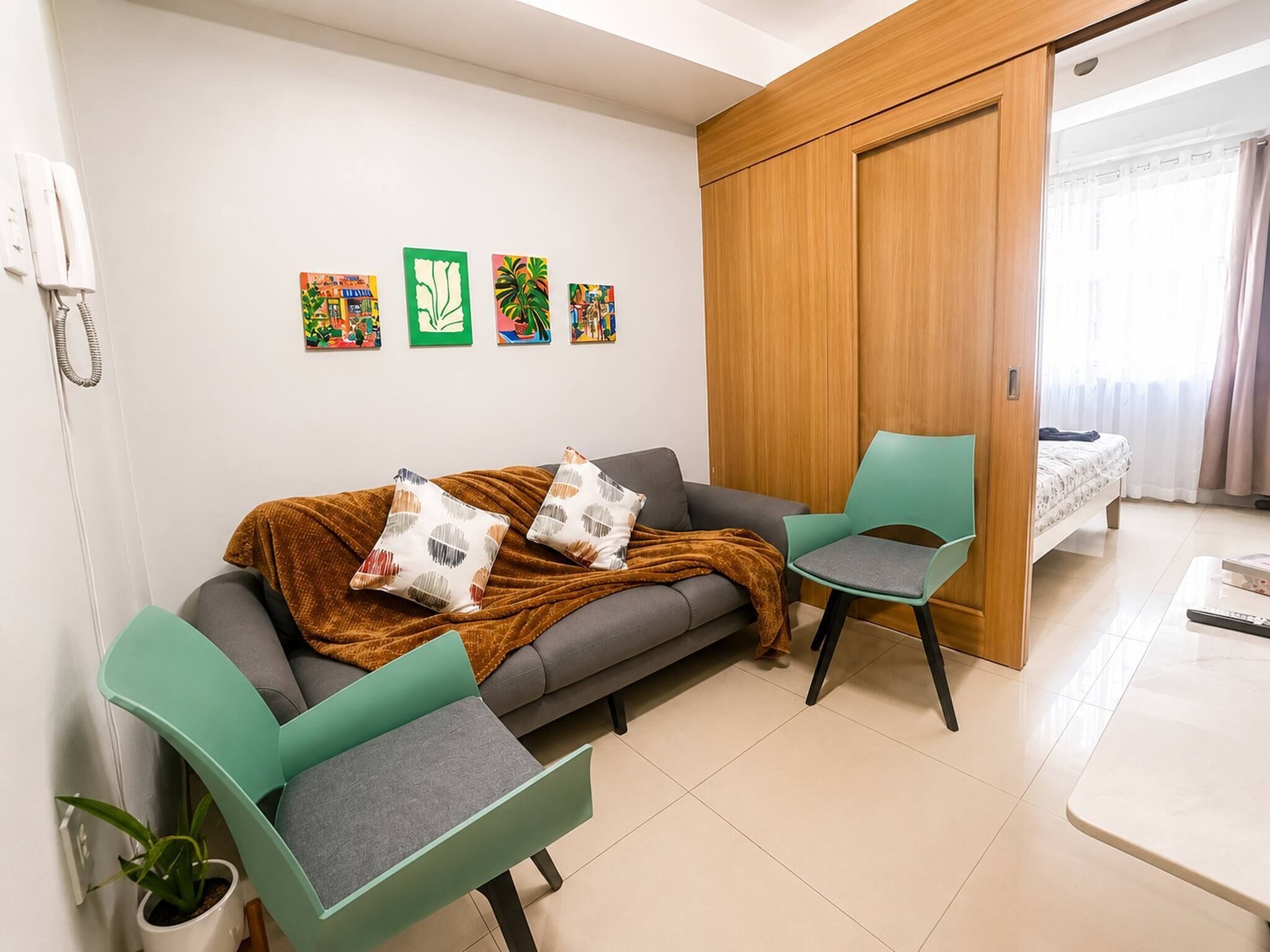 A modern living room with a sofa, two chairs, a wooden wardrobe, and a bed in the background.