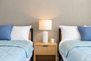 Ett hotellrum med två sängar, var och en med ett blått överkast och vita kuddar, ett nattduksbord med en lampa och en mugg samt en vägg med en intressant yta.