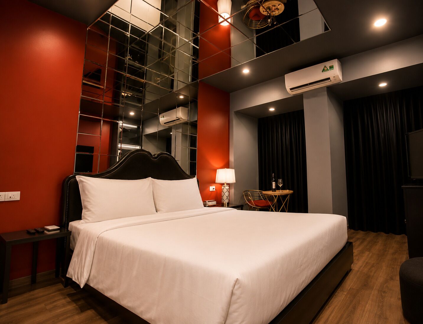 A hotel room with a large bed, a bedside table, a lamp, a chair, an air conditioning unit, and a television.