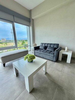 A modern living room with a grey sofa, a white coffee table with a green plant, and a view of a green landscape through large windows. - Beautiful, relaxing and convenient in Nuevo Vallarta! (Nuevo Nayarit)