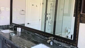 A bathroom with a large mirror, two sinks, a shower, and a marble countertop.