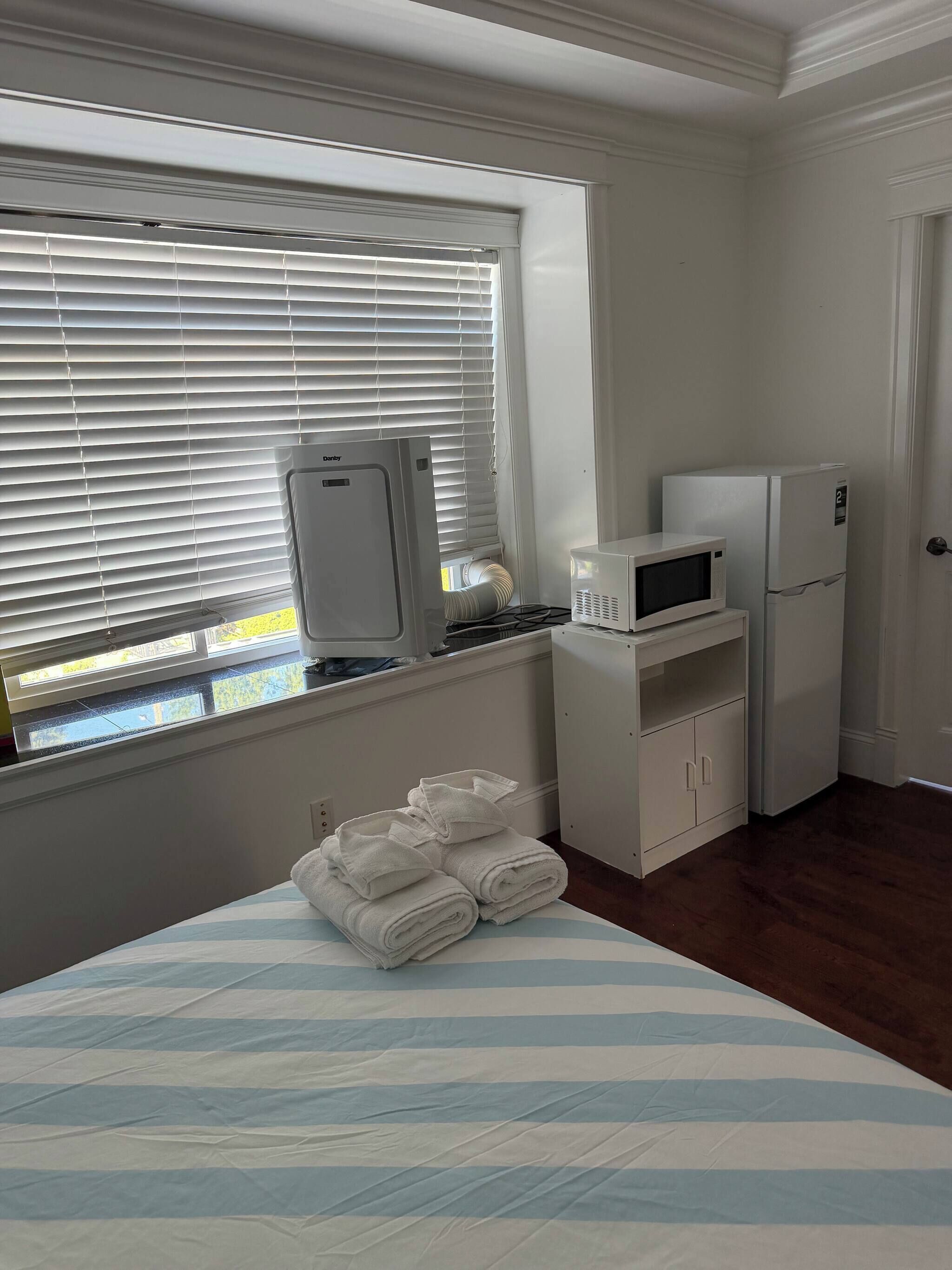 A hotel room with a bed, a window with blinds, a microwave, a refrigerator, and a stack of towels.