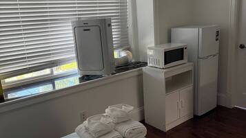 A hotel room with a bed, a window with blinds, a microwave, a refrigerator, and a stack of towels.