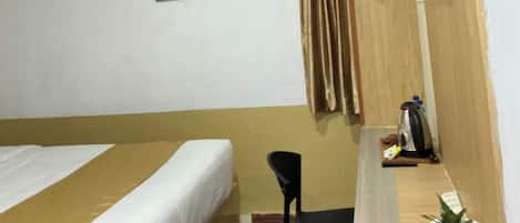 Quarto de hotel com cama, escrivaninha, cadeira, televisão fixada na parede e uma pequena planta em vaso.