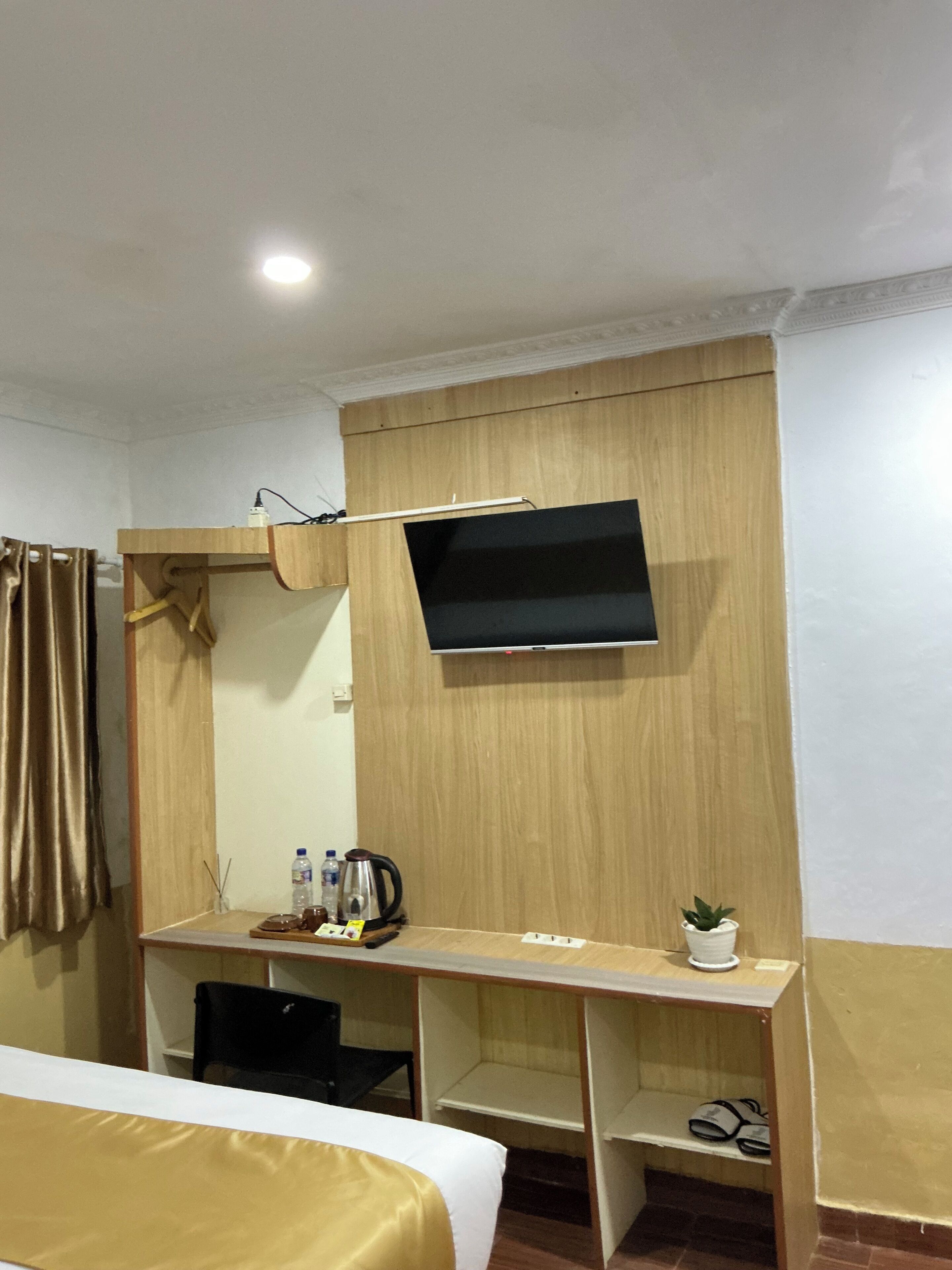 A hotel room with a bed, a desk with a flat-screen TV, a small potted plant, and a shelf with slippers.