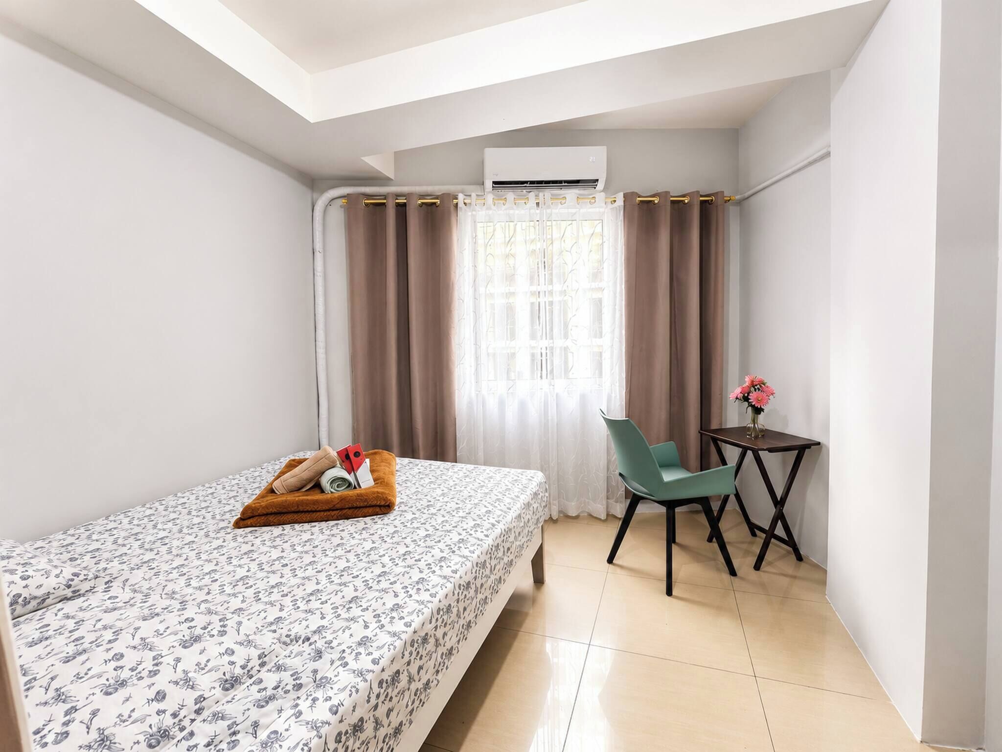 Une chambre d’hôtel de taille modeste, comprenant un lit, une petite table avec un vase de fleurs, une chaise et un appareil de climatisation.