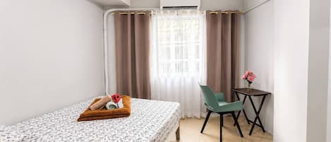 Une chambre d’hôtel de taille modeste, comprenant un lit, une petite table avec un vase de fleurs, une chaise et un appareil de climatisation.