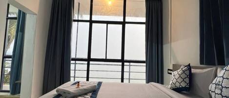 Ein Hotelzimmer mit einem großen Bett, einem fensterbelaubten Fenster, einem Nachttisch und Blick nach draußen.