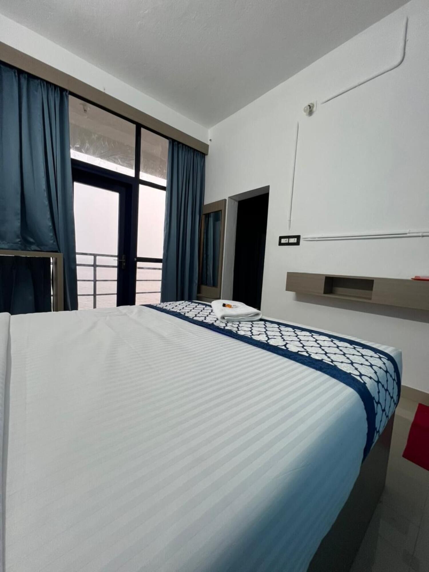 A hotel room with a large bed, a window with curtains, a door, and a wall-mounted air conditioning unit.