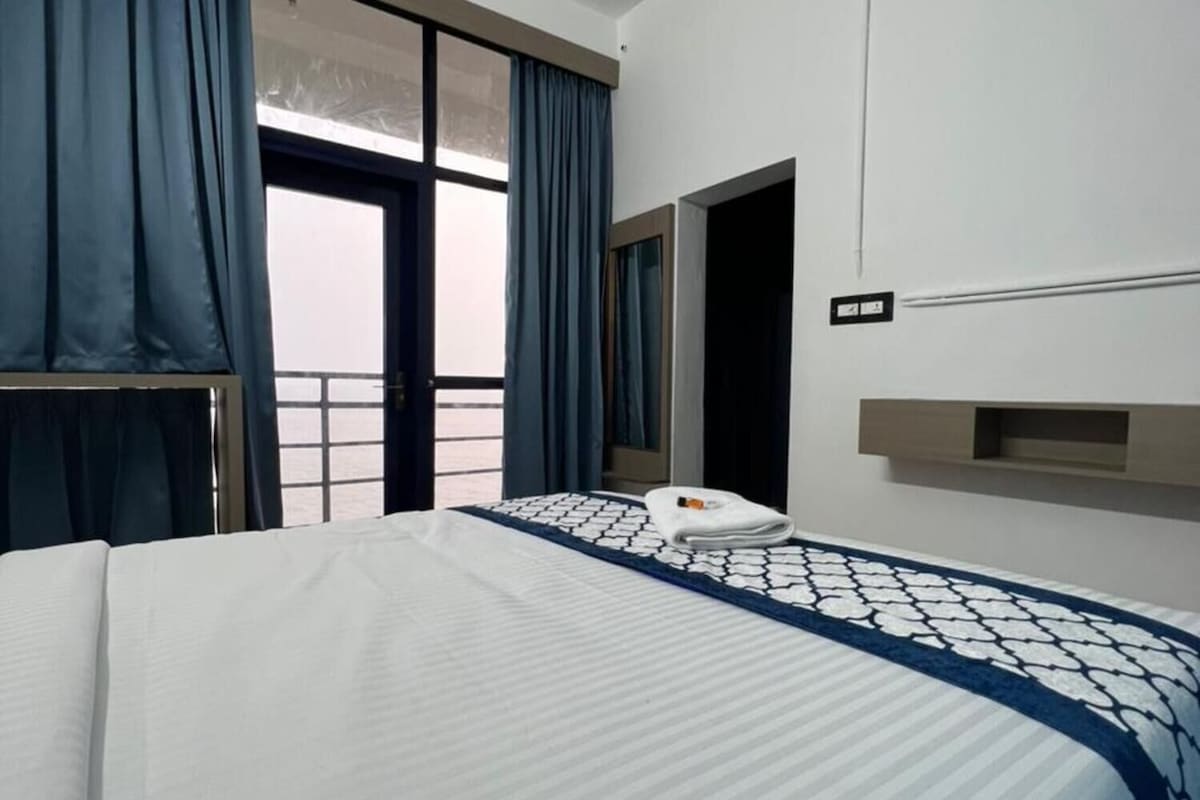 Habitación de hotel con una cama grande, una ventana con cortinas, una mesita de noche y un aplique de pared.