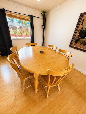 A round wooden dining table with eight chairs, a black curtain, a potted plant, and a framed painting on the wall.