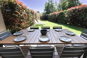 An outdoor dining area with a wooden table set for six, surrounded by chairs, and a hedge-lined pathway leading to a white lounge chair. - Villa - 3 bedrooms - 800 meters from Cala Rossa Beach (Lecci)