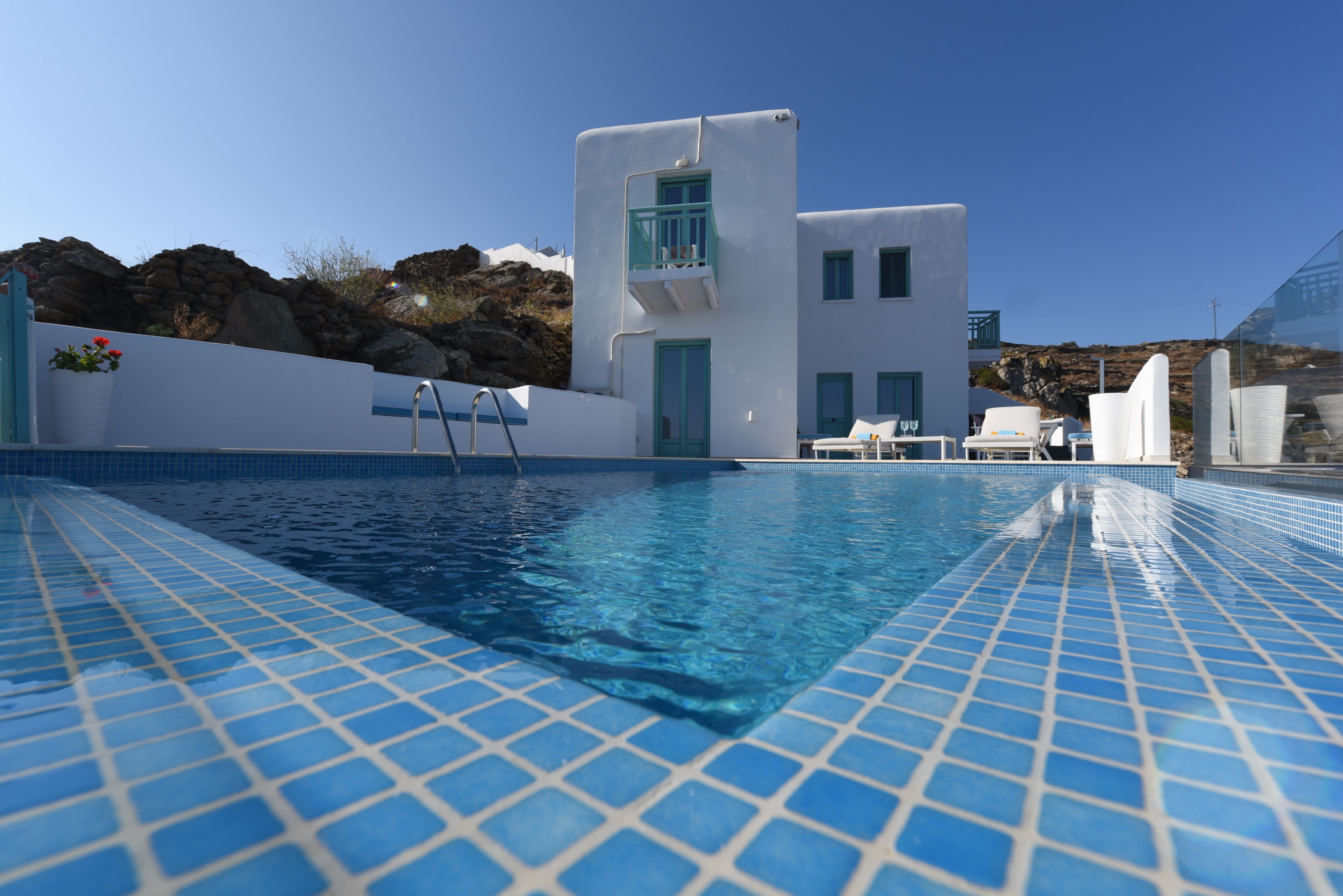 Ein Swimmingpool mit klaren blauen Fliesen, ein weißes Gebäude mit Balkon und ein felsiger Hügel im Hintergrund.