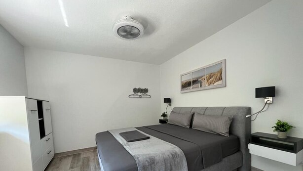 Ein modernes Schlafzimmer mit einem Bett, Nachttischen, einem an der Wand montierten Fernseher und einem Deckenventilator.