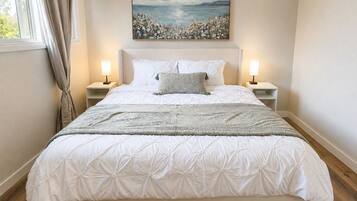 A bedroom with a large bed, two bedside lamps, a painting above the bed, and a window with curtains.