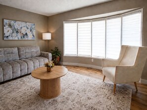 Ein Wohnzimmer mit einer Couch, einem Sessel, einem runden Couchtisch und einem großen Fenster mit Jalousien.