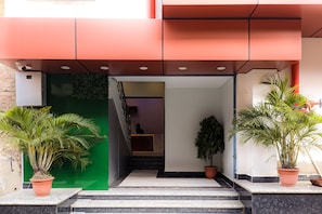 A hotel entrance with a reception desk, a staircase leading to the interior, and potted plants.