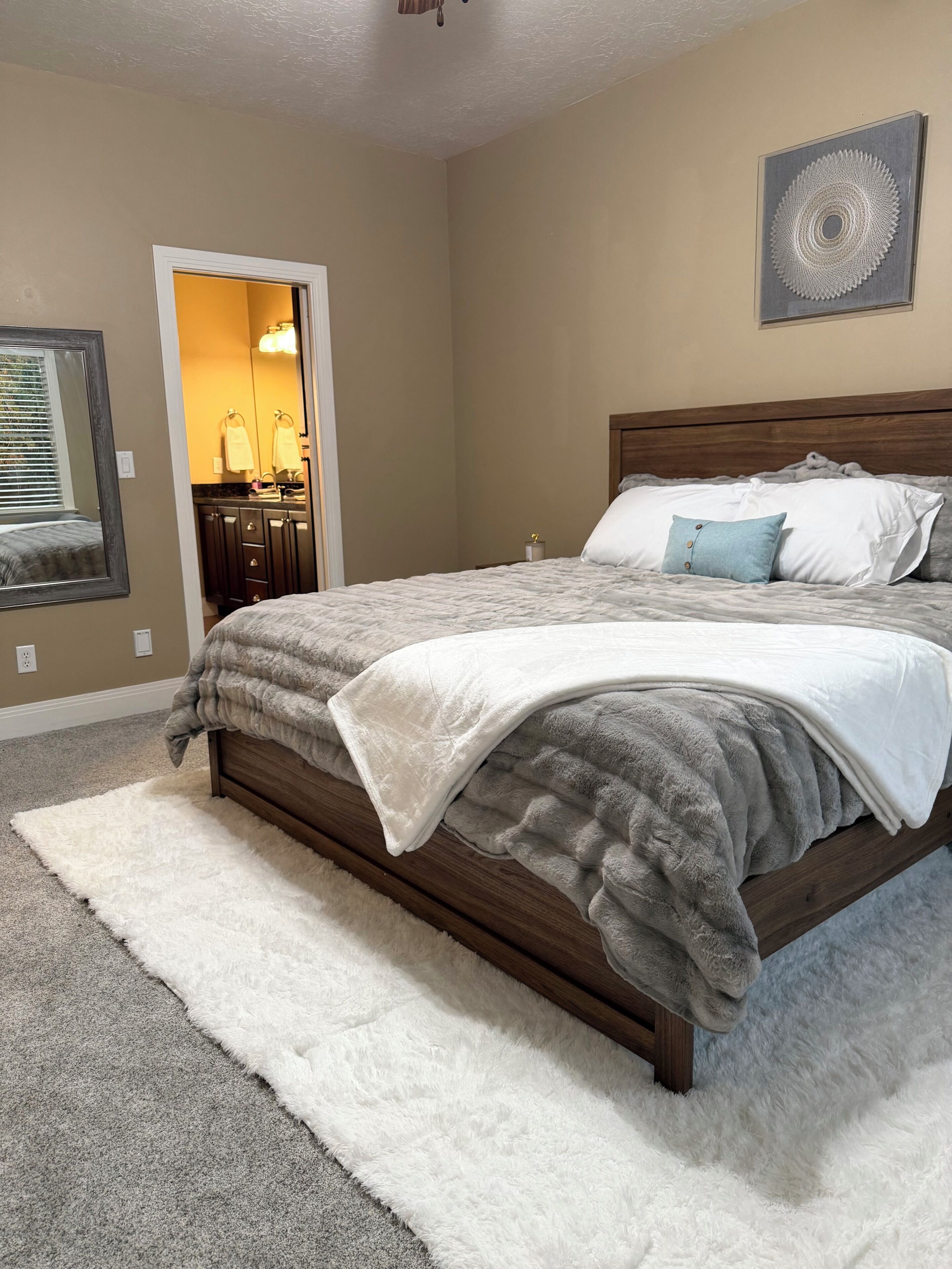A bedroom with a large bed, a wooden headboard, a white fluffy rug, and a wall-mounted artwork.