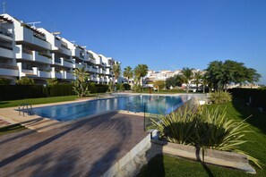 A swimming pool with a wooden deck, surrounded by well-maintained grass and palm trees, with a white building in the background. - Belvilla by OYO Dolce Vita (Orihuela Costa)