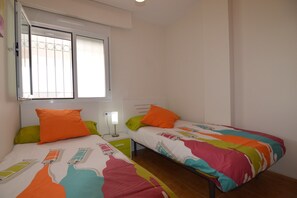 A room with two beds, each with colorful bedding and pillows, a window with blinds, a bedside table with a lamp, and a radiator. - Belvilla by OYO Dolce Vita (Orihuela Costa)