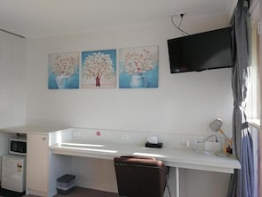 A room with a desk, a chair, a flat-screen TV mounted on the wall, and three paintings of trees in vases.