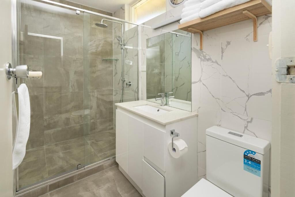 Un baño moderno con ducha envuelta en vidrio, un lavamanos blanco con espejo y un inodoro con etiqueta de clasificación de agua.