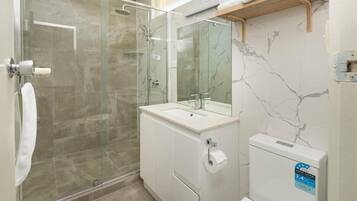 Un baño moderno con ducha envuelta en vidrio, un lavamanos blanco con espejo y un inodoro con etiqueta de clasificación de agua.