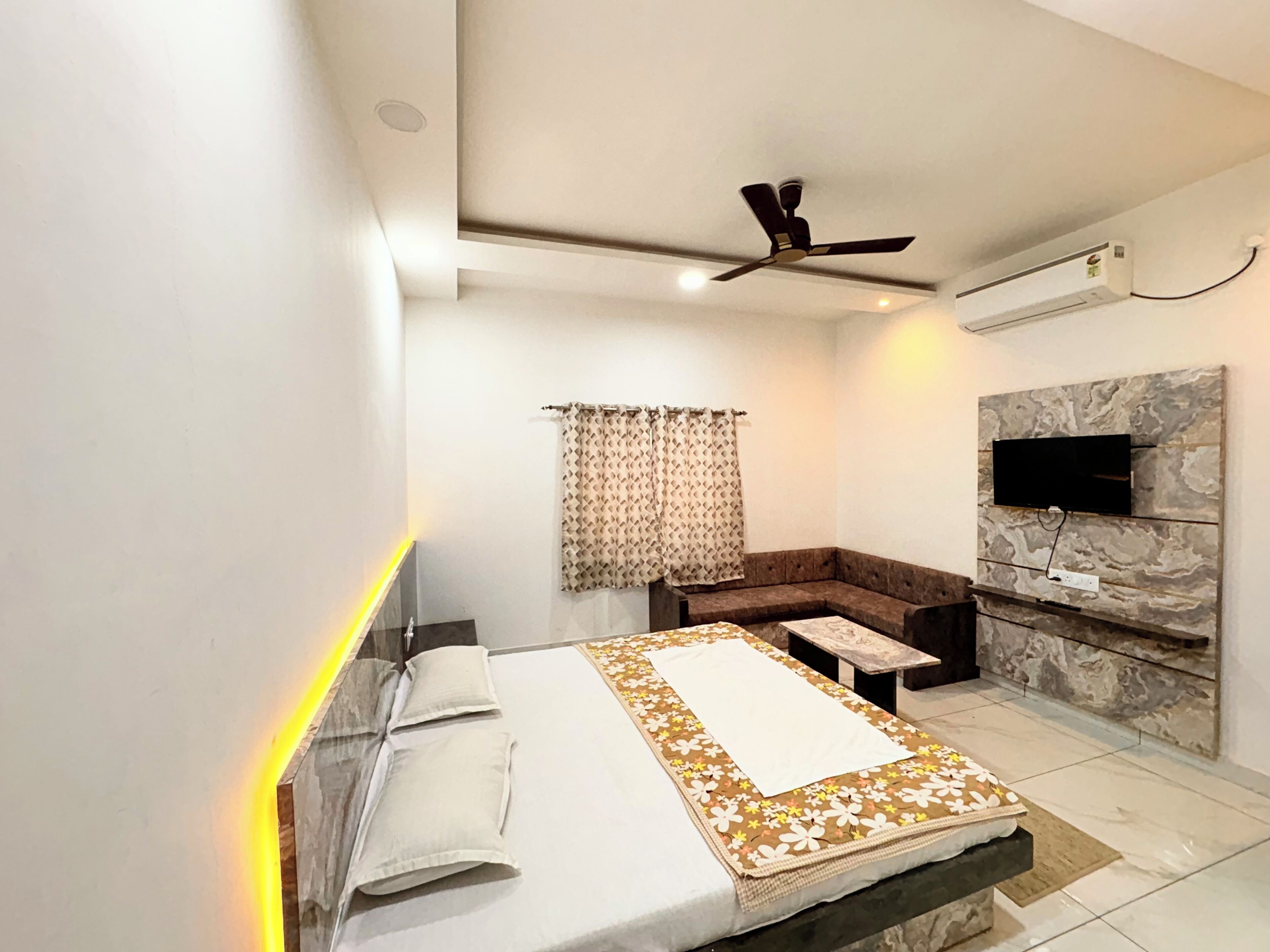A modern hotel room with a bed, a sofa, a flat-screen TV mounted on a stone wall, and a ceiling fan.