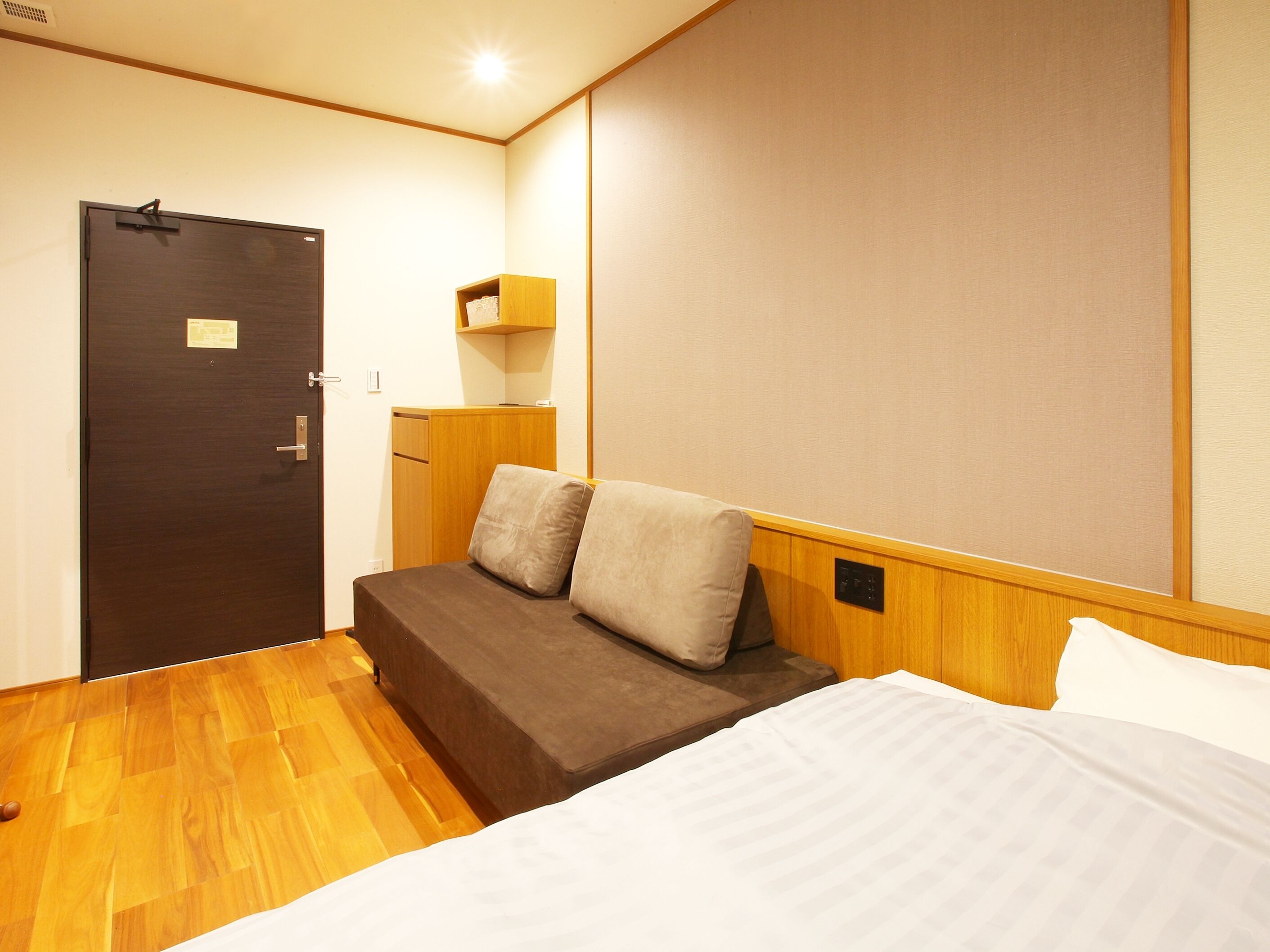 A compact hotel room with a bed, a sofa, a wooden wardrobe, and a small shelf.