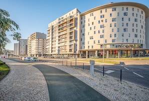 Un immeuble à plusieurs étages, avec un espace commercial au rez-de-chaussée, un trottoir pavé et des voitures garées.