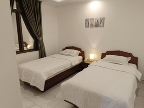A hotel room with two single beds, a nightstand with a lamp, a window with curtains, and a wall with three framed pictures. - Spacious 3-bedroom apartment with AC in serene Jogja (Jogja)