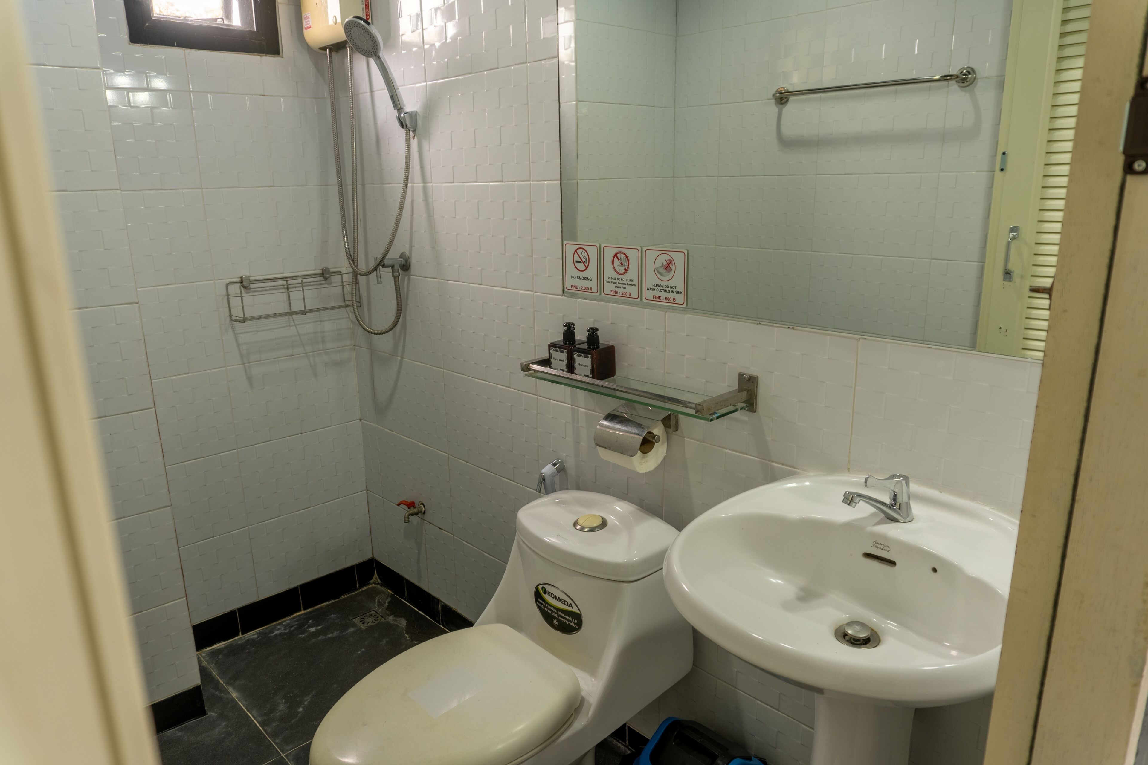 Une petite salle de bain équipée de toilettes, d’un lavabo et d’une douche. Des panneaux indiquent qu’il est interdit de fumer et de laver du linge dans le lavabo.