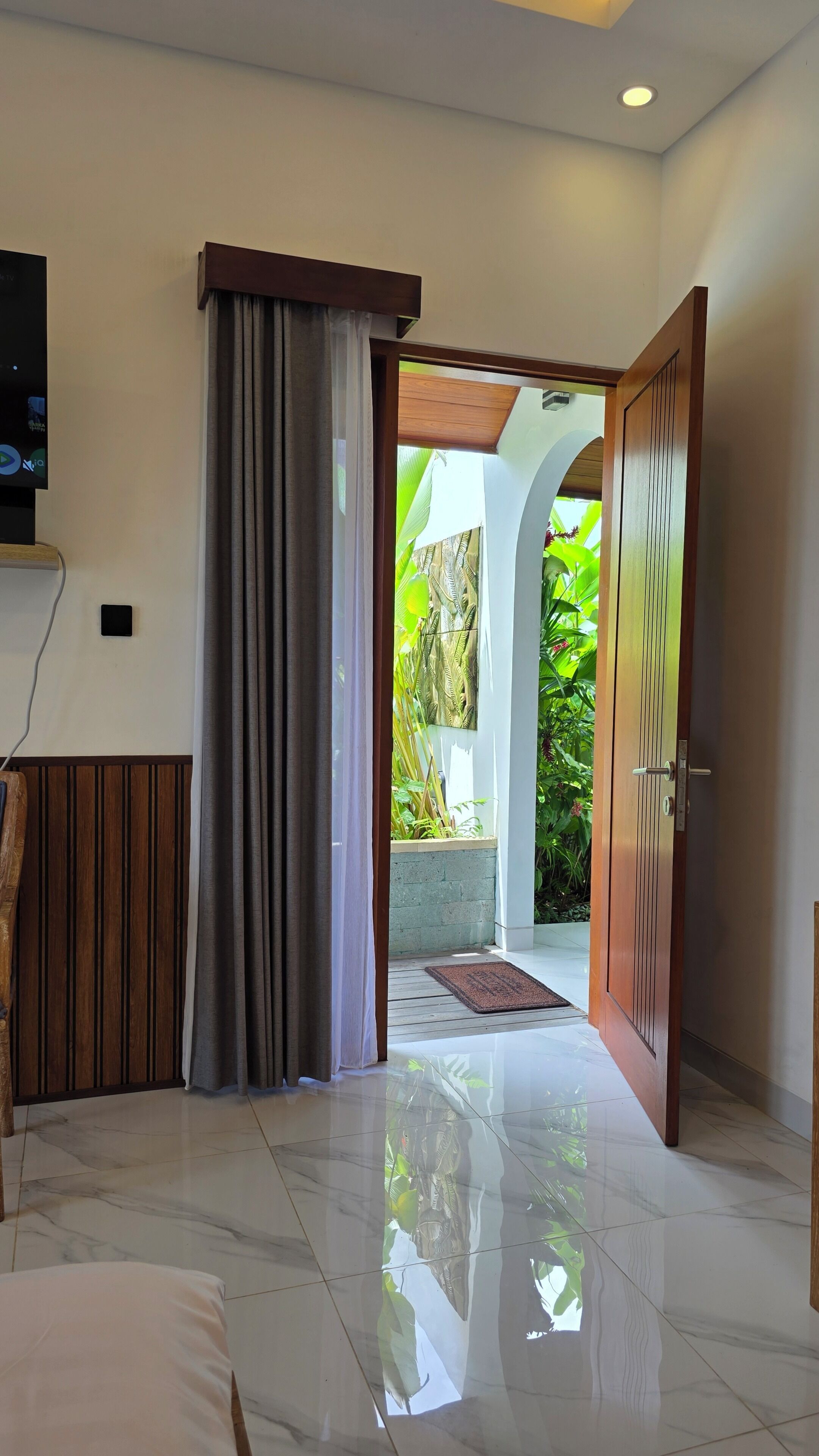 Habitación de hotel con puerta de madera, televisor y vista a un jardín desde una puerta abierta.