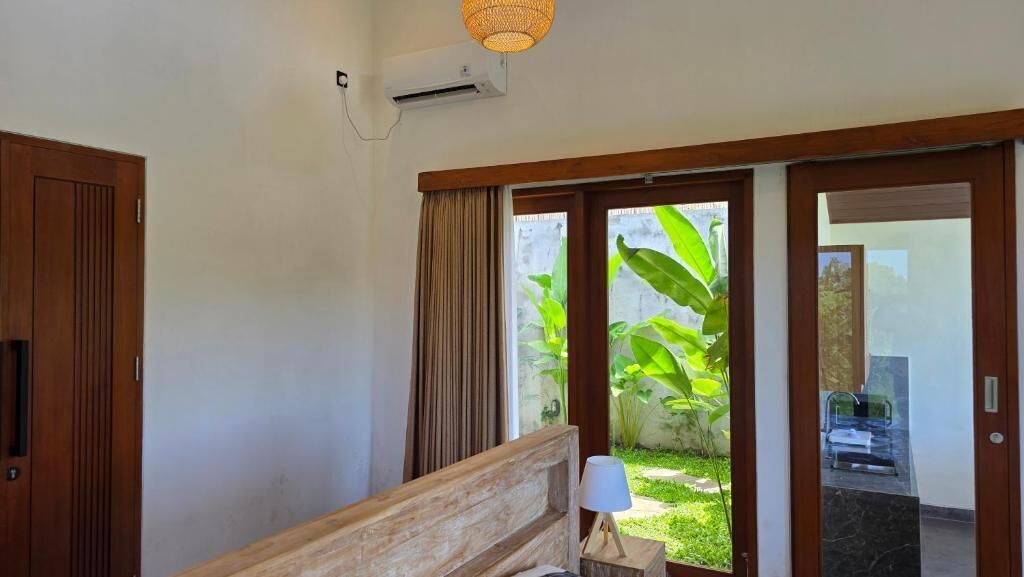 A hotel room with a wooden bed, a bedside lamp, a window with curtains, and an air conditioning unit.