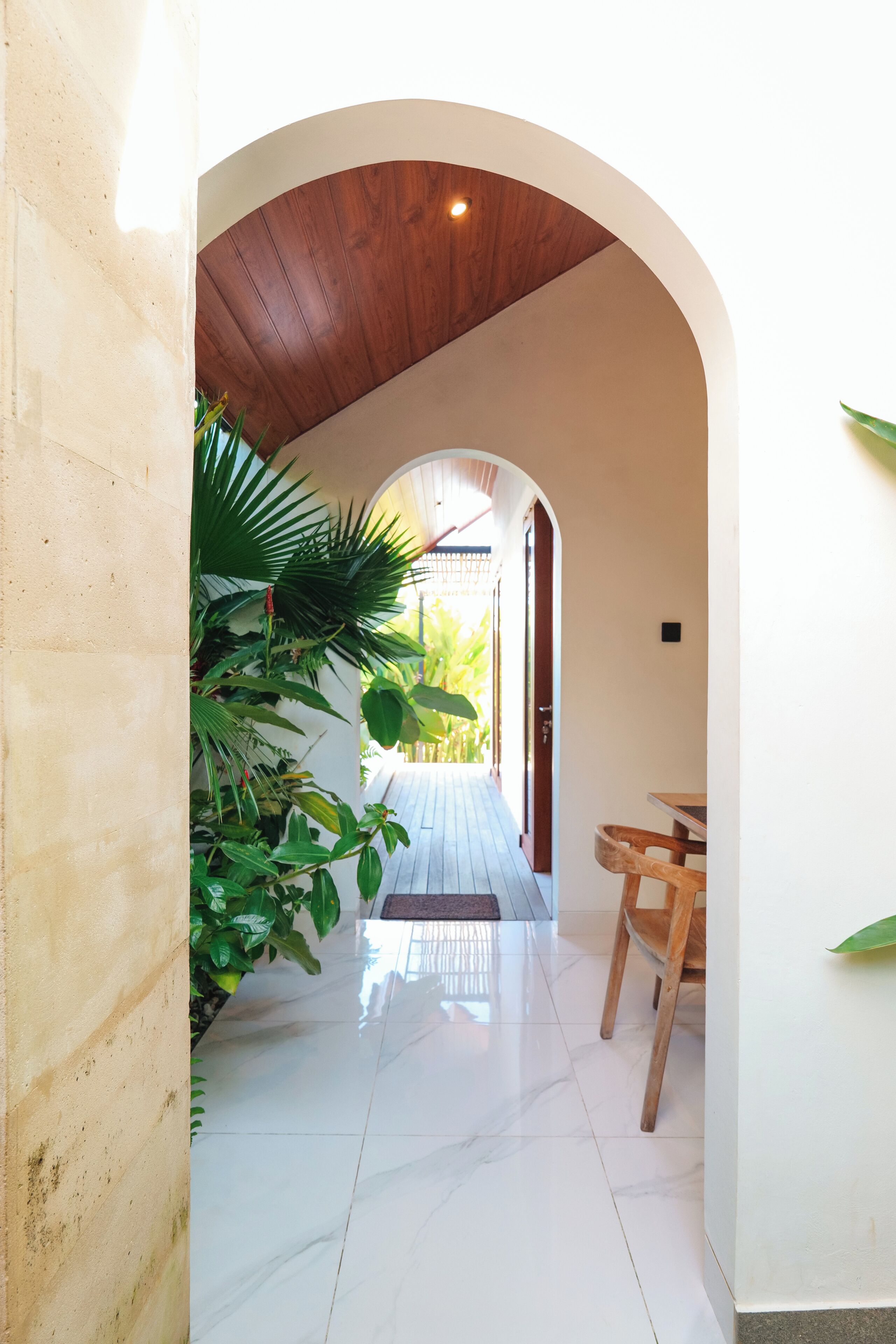 Um corredor com teto de madeira, chão de azulejos e vista para um jardim através de um arco aberto.