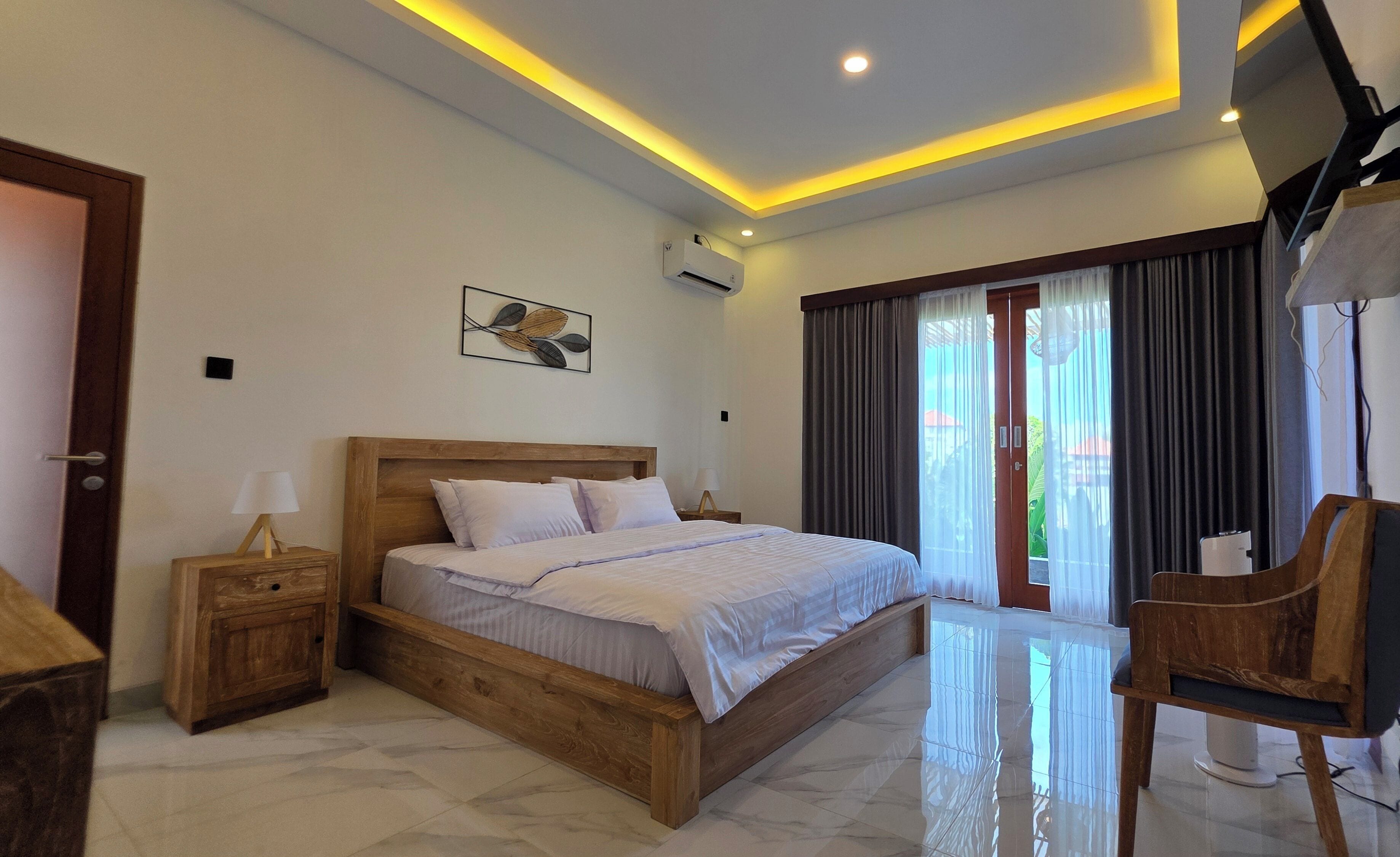 Quarto de hotel com cama de madeira, mesa de cabeceira, uma cadeira, televisão e uma janela grande com cortinas.