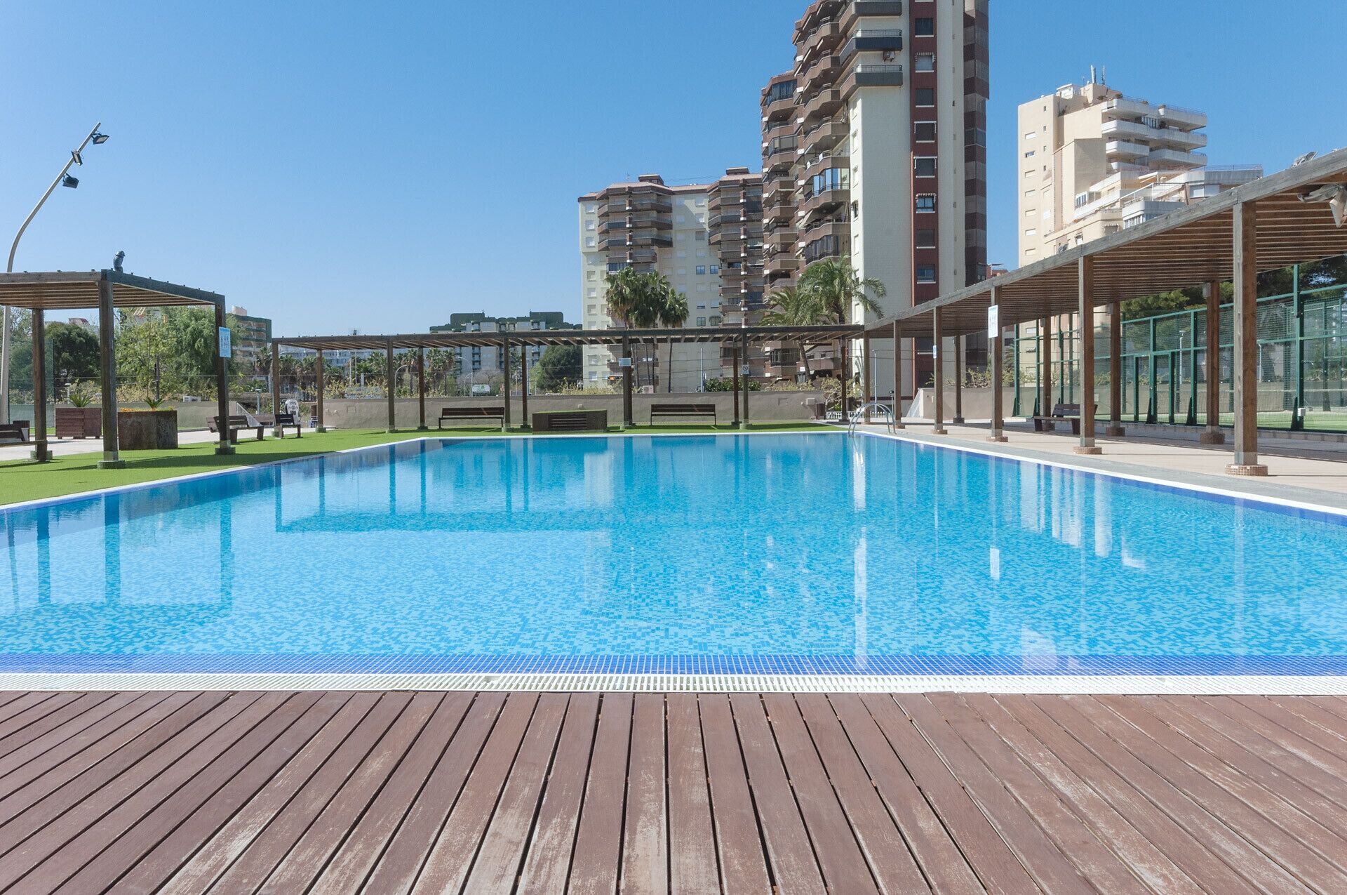 A swimming pool with a wooden deck, surrounded by buildings and a clear sky.