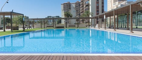 A swimming pool with a wooden deck, surrounded by buildings and a clear sky.