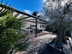 A covered outdoor patio with a grill, dining table, and chairs, adjacent to a white building with a window and door. - Beachfront Retreat With Private Canopy (Kamperland)