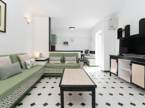 A compact living space with a green sofa, a wooden table, and a kitchen area with a TV and microwave. - Vivelorural Campo de Golf 76 (San Bartolomé de Tirajana)