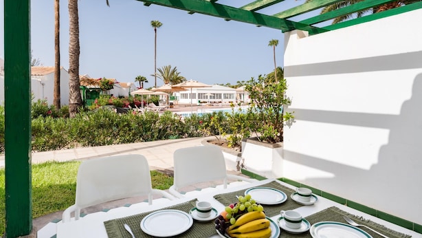 An outdoor dining area with a table set for two, a pool in the background, and a view of a building with a tiled roof. - Vivelorural Campo de Golf 76 (San Bartolomé de Tirajana)