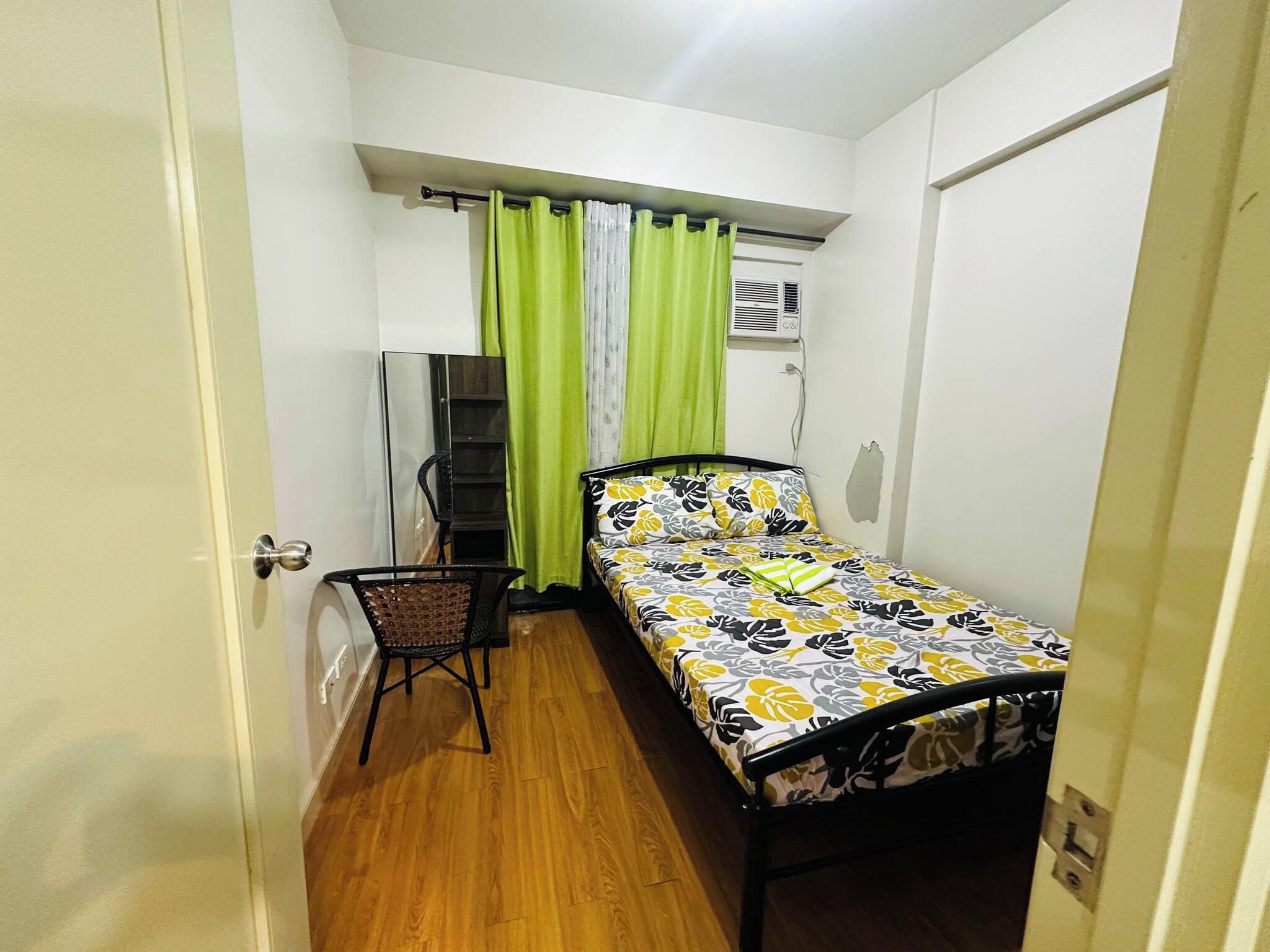 A small, single-bed room with a wooden floor, a chair, a desk, and a window with curtains.