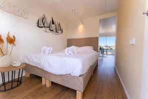 A bedroom with a bed, a round wooden table with a vase and dried plants, and a wall-mounted artwork.