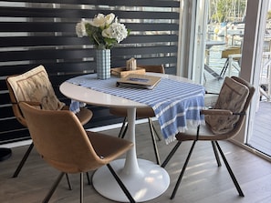 A dining area with a round table, two chairs, a vase with flowers, and a view of boats through a window.